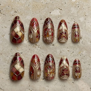 May include: A set of ten press-on nails featuring a stained glass design with red, cream, and gold geometric patterns. Each nail has a glossy finish with raised gold lines outlining the intricate shapes, arranged in two rows of five varying sizes.