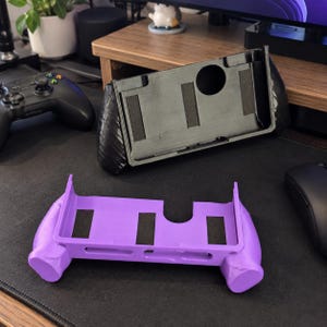 May include: Two handheld gaming console grips, one in solid black and one in solid purple. Each grip features a rectangular frame with integrated side handles, a circular cutout on the back, and three rectangular foam pads on the interior surface.