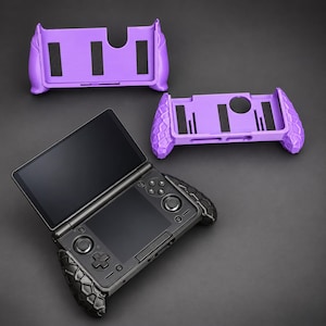 Puede incluir: Una consola de juegos portátil negra con diseño de doble pantalla y controles integrados, acompañada de dos fundas protectoras moradas. Las fundas presentan un patrón texturizado de panal en los agarres laterales y tiras de acolchado negro.