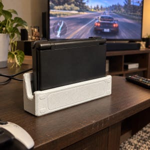 May include: A black handheld gaming console rests vertically in a light gray, rectangular, textured plastic charging dock. The dock features a recessed central panel with small rectangular indentations at each corner, providing a stable base for the device.