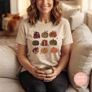 Peut inclure: Un t-shirt beige à manches courtes orné d'un imprimé graphique représentant neuf citrouilles disposées en grille de trois par trois. Chaque citrouille présente un motif unique, incluant des textures florales, à carreaux, rayées et unies.