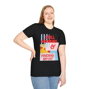 May include: A black short-sleeved t-shirt featuring a graphic design with the red text STILL GROWING & FINDING MY FIT. The design includes various rectangular patterns in blue, yellow, pink, and white with stripes, dots, and floral motifs.