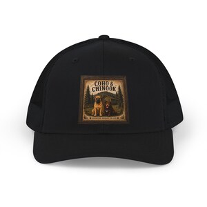 May include: A black trucker hat featuring a square graphic print on the front. The print shows two dogs in front of a cabin and pine trees, with the text COHO & CHINOOK and ADVENTURE BUDDIES FOR LIFE. The hat has a solid front panel and mesh back.