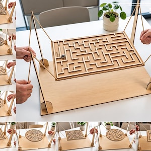 Könnte beinhalten: Ein hölzernes Tisch-Labyrinthspiel mit einem rechteckigen Brett, das an Schnüren befestigt ist, die an vier vertikalen Stützen hängen. Das Labyrinth enthält eine Metallkugel und besteht aus hellem Holz. Der Text 3MM ist sichtbar.