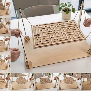 Pode incluir: Um jogo de labirinto de mesa em madeira com um tabuleiro retangular suspenso por cordas a partir de quatro suportes verticais. O jogo inclui uma esfera de metal que navega pelos caminhos do labirinto ao inclinar o tabuleiro à mão.