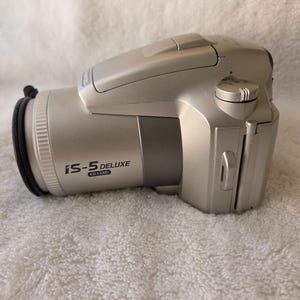 May include: A silver Olympus iS-5 Deluxe camera with an ED lens. The camera body features a prominent lens barrel, a top-mounted flash compartment, and a control dial on the side. The model name iS-5 DELUXE is printed clearly on the side of the lens.