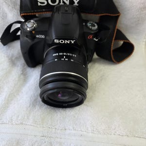 May include: A black Sony Alpha 230 DSLR camera with a 3.5-5.6/18-55 SAM lens attached. The camera features a black neck strap with orange trim and the Sony logo printed in white on the strap and the camera body. The lens barrel shows focal length markings.