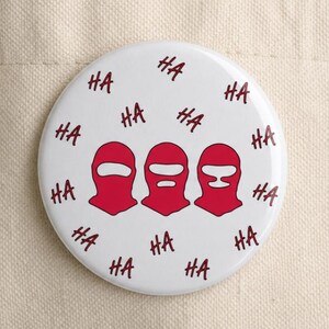 May include: A circular white pinback button featuring three red balaclava-style masks in a row, surrounded by the repeated dark red text "HA" scattered across the surface, attached to a plain beige canvas tote bag.