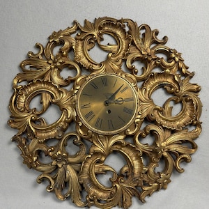 May include: A circular wall clock with a metallic gold finish, featuring an intricate, ornate frame with scrolling leaf and floral motifs. The central clock face has a smooth gold surface with black Roman numerals and two thin black hands.
