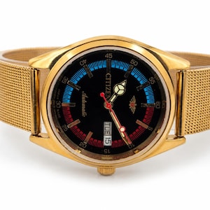 Op de afbeelding: Een goudkleurig Citizen automatisch horloge met een zwarte wijzerplaat, voorzien van een metalen mesh-band, lichtgevende wijzers, een rode secondewijzer en een dag-datumvenster op de drie-uurpositie. De wijzerplaat heeft blauwe en rode accenten.