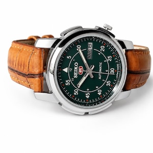May include: A Seiko 5 automatic wristwatch with a round silver-tone stainless steel case and a dark green dial. The dial features white Arabic numerals, luminous hands, and a day-date window. It is attached to a textured tan leather strap.