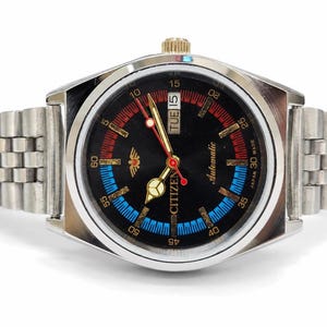 May include: A silver-toned Citizen automatic wristwatch with a black dial, featuring gold-tone hands, a red second hand, and a day-date window at the three o'clock position. The dial includes red and blue accents and a metal link bracelet.