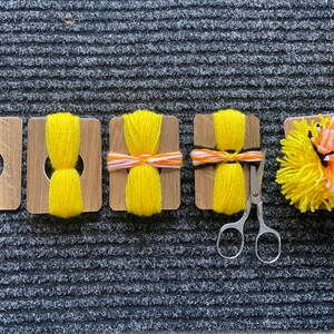 May include: A set of five wooden pom-pom makers showing the crafting process. One is empty, three are wrapped with yellow, orange, and white yarn, and one is a finished yellow, orange, and white pom-pom. Small metal scissors are included.