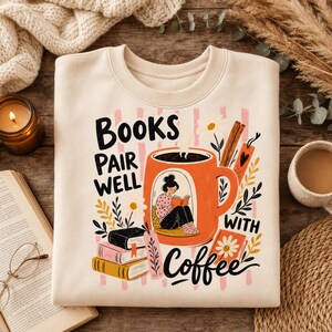 Può includere: Una felpa girocollo color crema con una stampa grafica di una tazza da caffè arancione contenente una persona che legge un libro. Il testo "BOOKS PAIR WELL WITH COFFEE" è affiancato da illustrazioni di libri impilati, fiori e bastoncini di cannella.