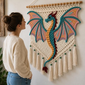 Puede incluir: Un tapiz de macramé color crema con un dragón tejido a ganchillo en tonos turquesa, amarillo y rosa. El dragón tiene alas grandes, una cola larga y una cresta roja. La pieza cuelga de una varilla de madera y está decorada con borlas.