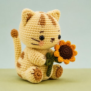 May include: A crocheted cat figurine in light beige yarn with brown tabby stripes on its head, back, and tail. The cat has black bead eyes, pink stitched cheeks, and holds a small crocheted sunflower with yellow petals and a dark brown center.