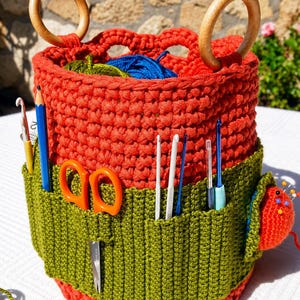 May include: A cylindrical orange crochet basket with two wooden ring handles, featuring an attached olive green exterior pocket panel. The basket contains blue and green yarn, while the pockets hold a blue pencil, orange scissors, and various crochet hooks.
