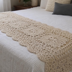 May include: A cream-colored rectangular crochet bed runner featuring a repeating floral motif pattern with a scalloped edge. The textile is draped horizontally across a white bedspread, showcasing intricate openwork stitching and a textured yarn finish.