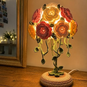 Puede incluir: Una lámpara de mesa con una pantalla hecha de flores de ganchillo en tonos rosa, amarillo y blanco. La base de la lámpara está cubierta con una funda circular de ganchillo en beige y marrón, con un tallo y hojas verdes de ganchillo.