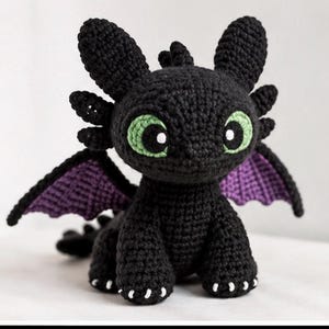 May include: A black crocheted dragon plush toy featuring large green eyes with black pupils, pointed ears, and small purple wings. The creature has a textured yarn surface, a rounded head, and small white stitches on its feet, resembling a mythical beast.