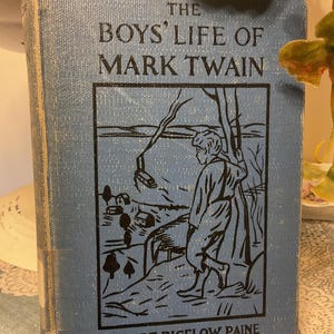May include: A light blue hardback book titled The Boys' Life of Mark Twain by Albert Bigelow Paine. The front cover features a black line drawing of a person standing near a tree overlooking a landscape with small houses and a boat on water.