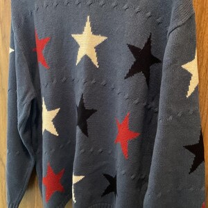 Könnte beinhalten: Ein langärmeliger Pullover mit Rundhalsausschnitt in staubigem Blau, der mit einem sich wiederholenden Muster aus fünfzackigen Sternen in Rot, Weiß und Schwarz versehen ist. Das Strickgewebe weist horizontale Reihen erhabener Punkte auf.