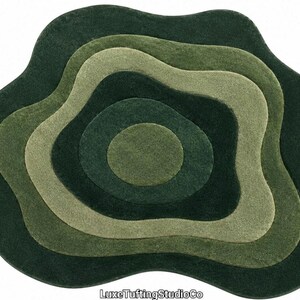 May include: An irregular, amoeba-shaped rug featuring concentric, wavy layers in varying shades of green, ranging from dark forest green to light sage. The rug has a soft, tufted texture and includes the text LuxeTuftingStudioCo at the bottom.