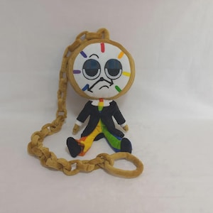 May include: A plush doll shaped like a clock with a round white face, large dark eyes, and a sad expression. It wears a black jacket over rainbow-colored pants and is attached to a long, thick, tan-colored plush chain that loops around the figure.