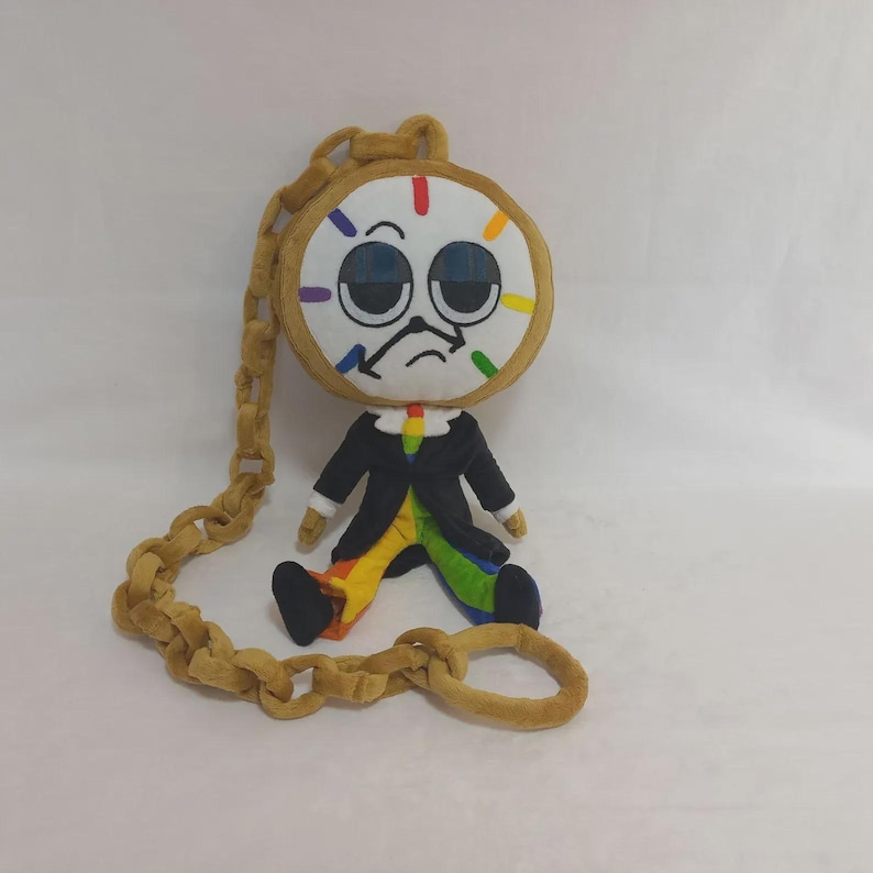 May include: A plush doll shaped like a clock with a round white face, large dark eyes, and a sad expression. It wears a black jacket over rainbow-colored pants and is attached to a long, thick, tan-colored plush chain that loops around the figure.