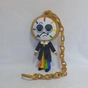 May include: A plush doll featuring a large round clock face with a bored expression, black suit jacket, and rainbow-colored pants. The doll is attached to a long, tan, soft-sculpted chain that loops around the head and extends to the floor.