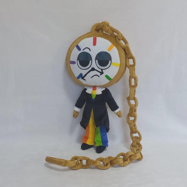 May include: A plush doll featuring a large round clock face with a bored expression, black suit jacket, and rainbow-colored pants. The doll is attached to a long, tan, soft-sculpted chain that loops around the head and extends to the floor.