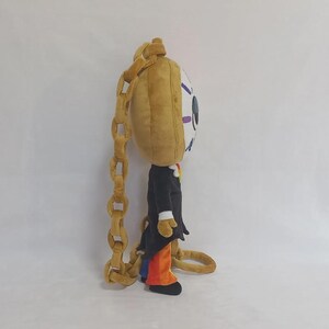 May include: A plush character toy featuring a large, round, tan head with a single blue eye and purple eyelashes. The figure wears a black tailcoat over orange trousers and is attached to a long, thick, tan plush chain that hangs down the back.