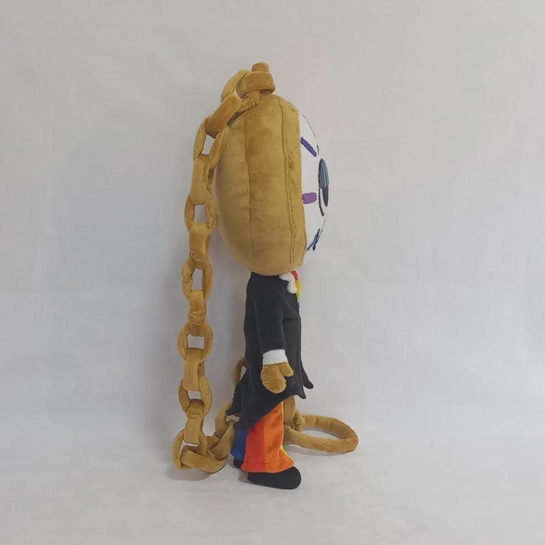 May include: A plush character toy featuring a large, round, tan head with a single blue eye and purple eyelashes. The figure wears a black tailcoat over orange trousers and is attached to a long, thick, tan plush chain that hangs down the back.