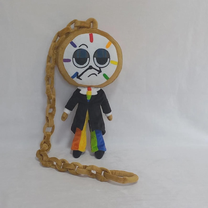 May include: A plush doll featuring a round clock face with a bored expression, colorful hour markers, and black hands. The doll wears a black jacket over a rainbow-striped shirt and pants, and is attached to a long, tan, plush chain link accessory.
