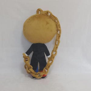 May include: A plush toy featuring a large, round, tan head and a black coat. The figure is encircled by a long, thick, tan plush chain that connects from the top of the head to the base, with small glimpses of blue and red fabric at the feet.
