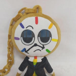 May include: A plush toy shaped like a clock with a round white face, large dark blue eyes, and a black clock hand mouth. It features a gold plush chain, a black suit jacket, and a colorful tie. The clock face has red, yellow, green, blue, and purple markers.