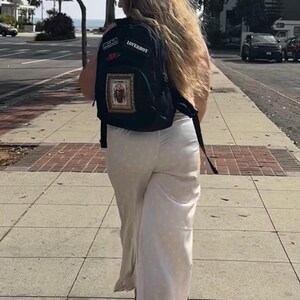 Puede incluir: Una mochila negra con un parche rectangular que muestra la ilustraci&oacute;n de un escarabajo, un parche m&aacute;s peque&ntilde;o con el texto LOVERBOY y un peque&ntilde;o parche rojo, combinada con pantalones largos de color claro con lunares hasta los tobillos.