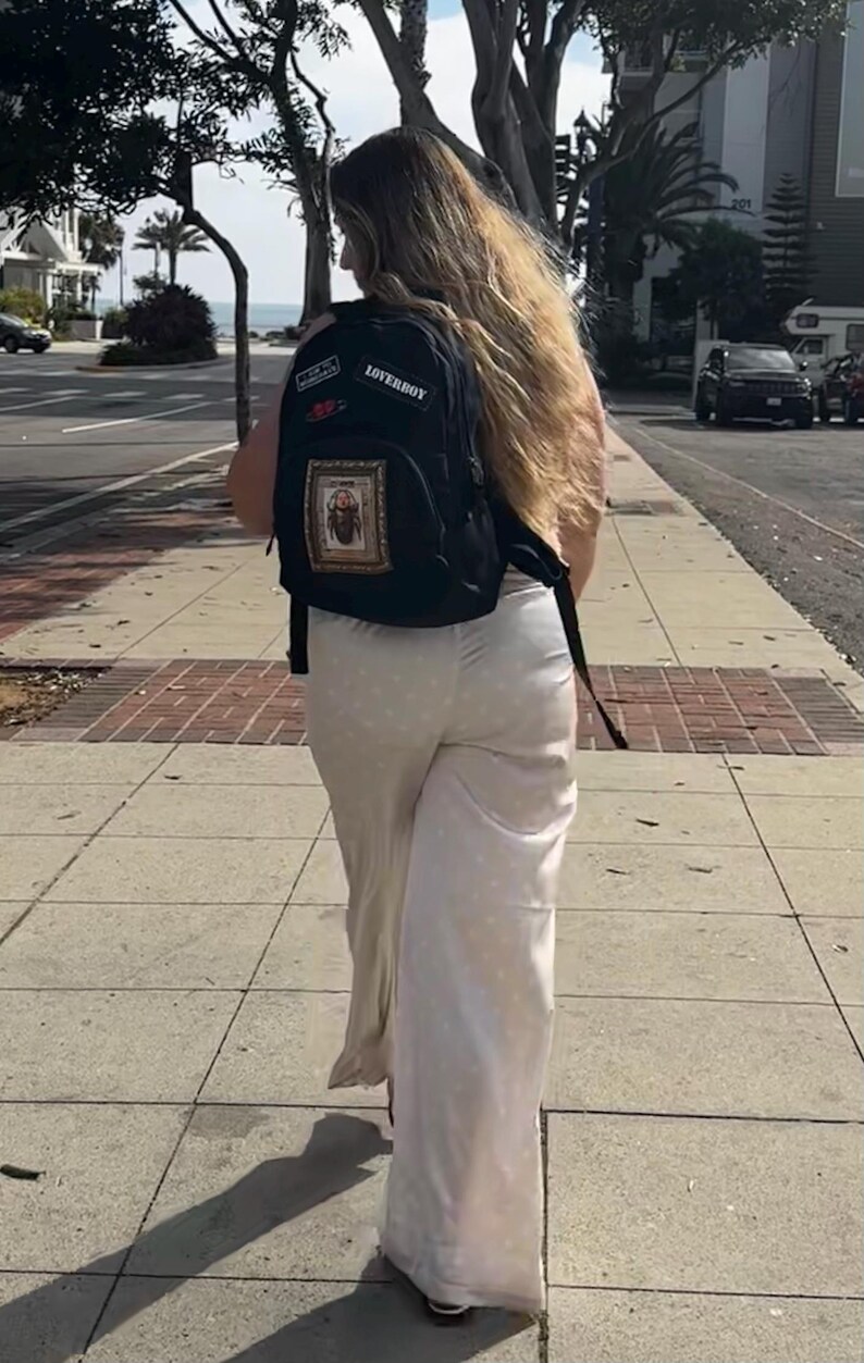 Puede incluir: Una mochila negra con un parche rectangular que muestra la ilustraci&oacute;n de un escarabajo, un parche m&aacute;s peque&ntilde;o con el texto LOVERBOY y un peque&ntilde;o parche rojo, combinada con pantalones largos de color claro con lunares hasta los tobillos.