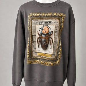 May include: A charcoal gray crewneck sweatshirt featuring a central graphic print of a beetle with a human face, set against a background of vintage newspaper clippings, all enclosed within a detailed, ornate gold-colored rectangular frame.