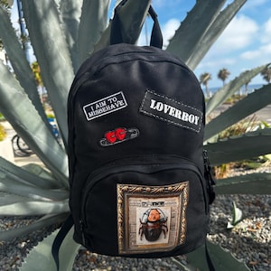 May include: A black backpack featuring a rectangular patch with the text I AIM TO MISBEHAVE, a rectangular patch with the text LOVERBOY, a safety pin with two red hearts, and a large rectangular patch depicting a beetle with a human face in a frame.