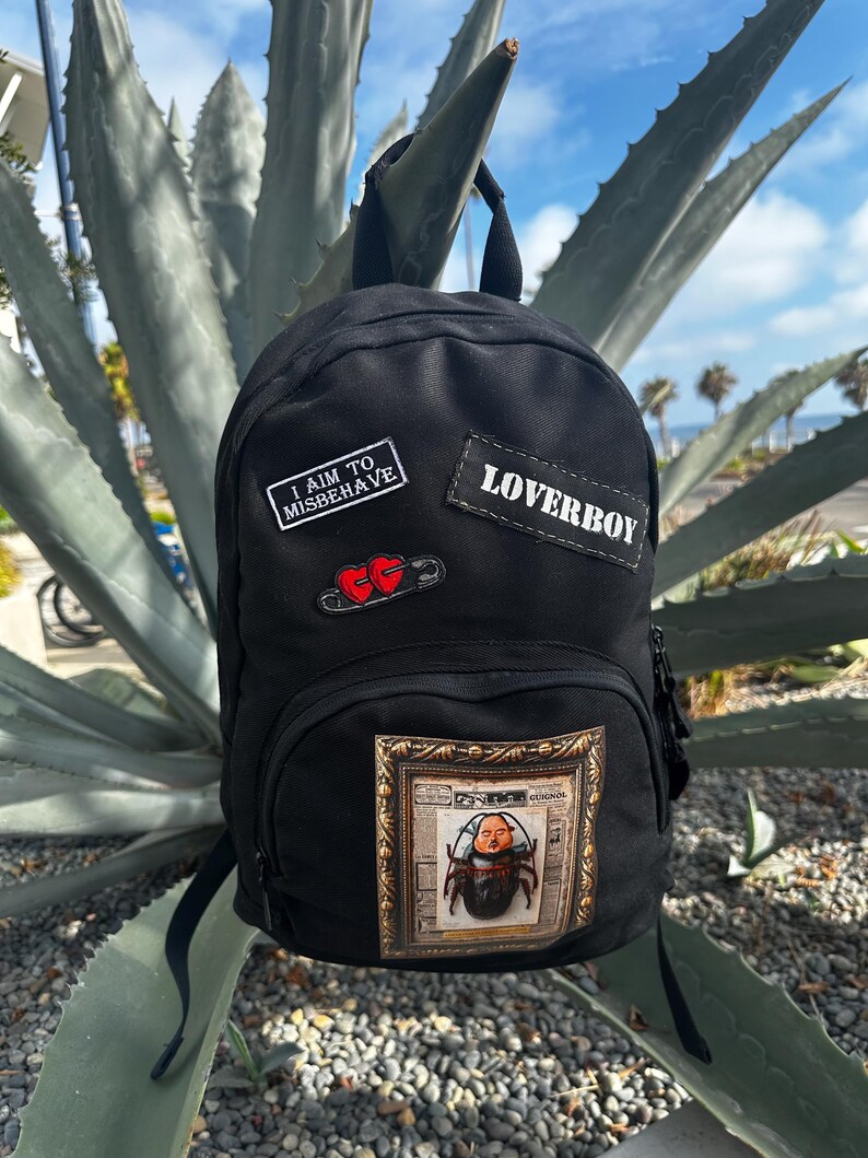 Puede incluir: Una mochila negra con un parche rectangular con el texto I AIM TO MISBEHAVE, un parche con el texto LOVERBOY, un imperdible con dos corazones rojos y un parche rectangular grande que representa un escarabajo con rostro humano en un marco.