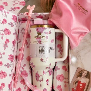 May include: A cream-colored 40 oz Stanley Quencher H2.0 tumbler with a pink rose pattern, a matching straw with a pink bow topper, a pink satin drawstring bag, and a pink ribbon, all presented in a floral-lined box with a promotional card.