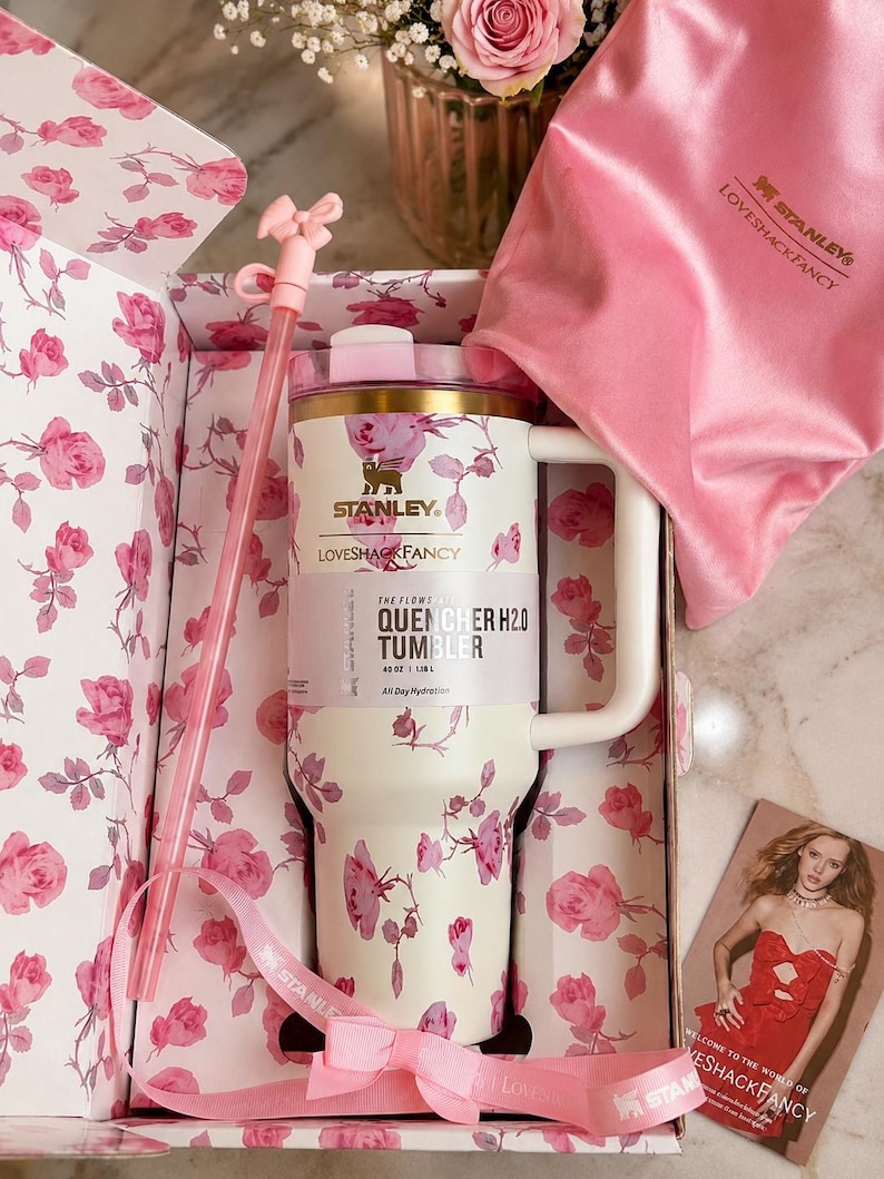 May include: A cream-colored 40 oz Stanley Quencher H2.0 tumbler with a pink rose pattern, a matching straw with a pink bow topper, a pink satin drawstring bag, and a pink ribbon, all presented in a floral-lined box with a promotional card.