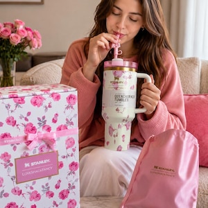 May include: A cream-colored Stanley Quencher H2O tumbler with a pink floral pattern, a gold rim, and a matching pink straw. The tumbler is accompanied by a white box with a pink floral print, a pink ribbon, and a pink satin drawstring bag.