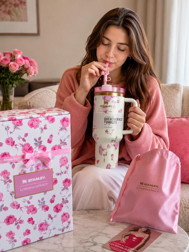 May include: A cream-colored Stanley Quencher H2O tumbler with a pink floral pattern, a gold rim, and a matching pink straw. The tumbler is accompanied by a white box with a pink floral print, a pink ribbon, and a pink satin drawstring bag.