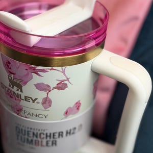 May include: A white Stanley Quencher H2.0 tumbler featuring a pink floral pattern, a gold-tone rim, and a white handle. The lid is clear with a pink border and a white rotating closure. The label reads Stanley, Fancy, and Quencher H2.0 Tumbler.
