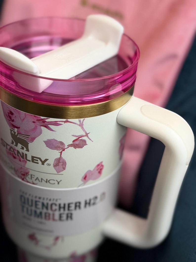 May include: A white Stanley Quencher H2.0 tumbler featuring a pink floral pattern, a gold-tone rim, and a white handle. The lid is clear with a pink border and a white rotating closure. The label reads Stanley, Fancy, and Quencher H2.0 Tumbler.