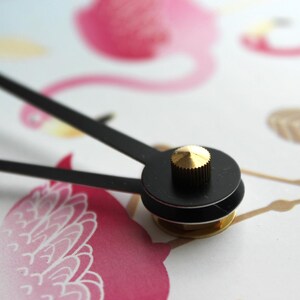 A close-up view of a clock face with a black dial and a gold-colored knob, set against a background with a pink flamingo and other abstract shapes.