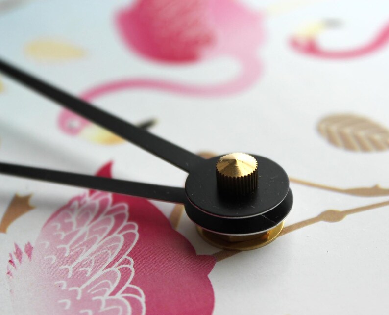 A close-up view of a clock face with a black dial and a gold-colored knob, set against a background with a pink flamingo and other abstract shapes.