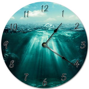 May include: A round clock with a blue sky and ocean scene. The clock face is divided into 12 hours, with the numbers 1 through 12 displayed in a white font. The clock hands are black. The image shows the ocean surface with a wave breaking and the water below the surface.